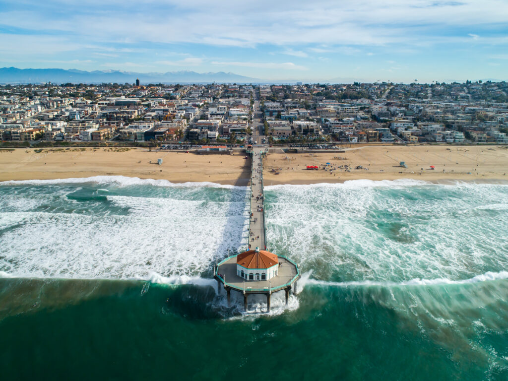 Manhattan Beach, California pier as seen from the ocean. Features pier with circular white building with orange roof and a long wooden boardwalk surrounded by ocean. The City buildings and residents are viewable in the background.