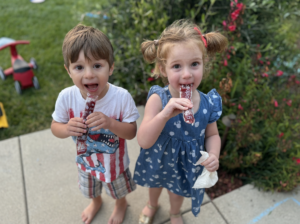 Two (2) young children - one girl and one boy - wearing fourth of July outfits and enjoying ice pops.